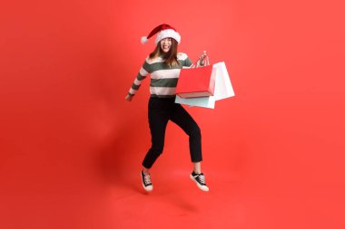 Kırmızı arka planda Noel Baba şapkalı ve alışveriş çantası taşıyan Asyalı bir kadın. Noel Baba! Noel Baba!.