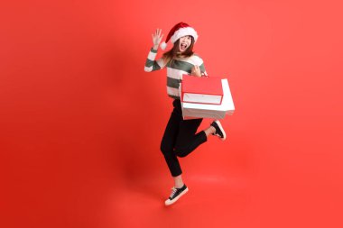 Kırmızı arka planda Noel Baba şapkalı ve alışveriş çantası taşıyan Asyalı bir kadın. Noel Baba! Noel Baba!.