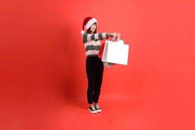 Kırmızı arka planda Noel Baba şapkalı ve alışveriş çantası taşıyan Asyalı bir kadın. Noel Baba! Noel Baba!.