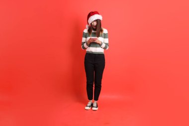 Kırmızı arka planda Noel Baba şapkalı ve cep telefonu tutan Asyalı bir kadın. Noel Baba! Noel Baba!.