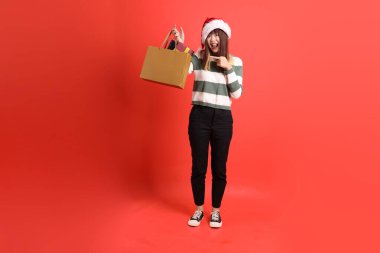Kırmızı arka planda Noel Baba şapkalı ve alışveriş çantası taşıyan Asyalı bir kadın. Noel Baba! Noel Baba!.