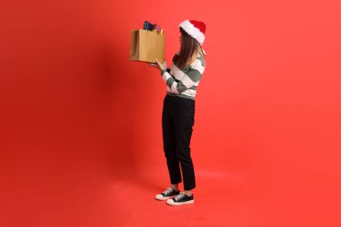 Kırmızı arka planda Noel Baba şapkalı ve alışveriş çantası taşıyan Asyalı bir kadın. Noel Baba! Noel Baba!.