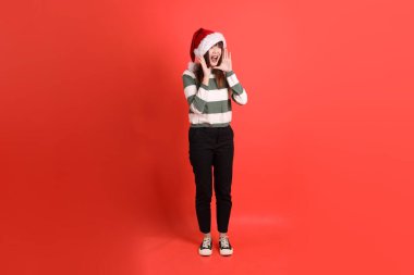 Noel ya da kışlık kıyafetler giyen Asyalı bir kadın. Noel Baba şapkası takmış, elinde kırmızı arka planda megafonla bağırıyor. Noel Baba! Noel Baba!.