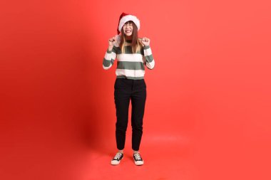 Noel ya da kışlık giysiler giyen Asyalı bir kadın Noel Baba şapkası takıyor. Heyecanlı, kırmızı arka planda izole edilmiş bir şekilde kutluyor. Noel Baba! Noel Baba!.
