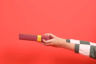 Yeşil çizgili süveter giyen bir el, kırmızı arka planda izole edilmiş bir hediye ya da hediye kutusu tutuyor. Mutlu Noeller..