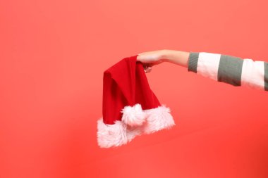 Kırmızı arka planda izole edilmiş, yeşil çizgili süveter giyen bir el Noel Baba şapkası tutuyor. Mutlu Noeller..