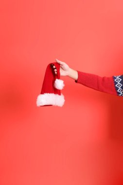 Kırmızı hırka giyen, kırmızı arka planda izole edilmiş bir el Santa Hat 'ı tutuyor. Mutlu Noeller..