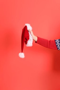 Kırmızı hırka giyen, kırmızı arka planda izole edilmiş bir el Santa Hat 'ı tutuyor. Mutlu Noeller..