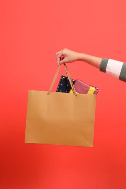 Yeşil çizgili süveter giyen, kırmızı arka planda izole edilmiş, alışveriş torbası tutan bir elin yakın çekimi. Mutlu Noeller..