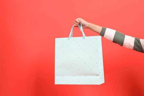 Крупный план руки в свитере с зеленой полосой, держащей сумку с покупками, на красном фоне. Счастливого Рождества!.