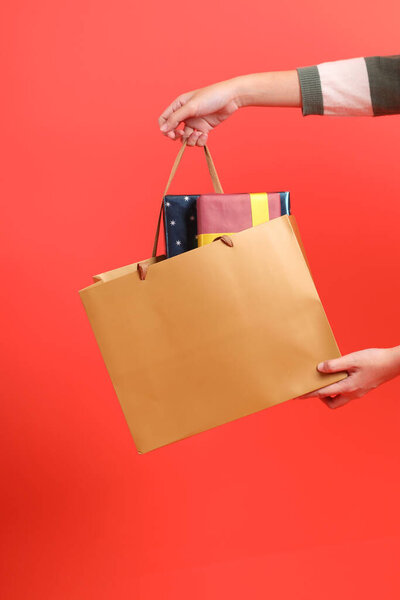 Крупный план руки в свитере с зеленой полосой, держащей сумку с покупками, на красном фоне. Счастливого Рождества!.