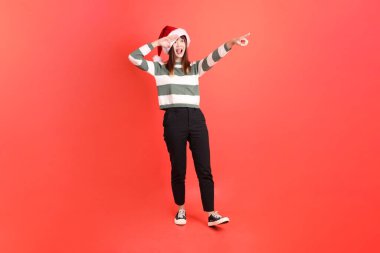 Noel ya da kışlık giysiler giyen Asyalı bir kadın. Noel Baba şapkası takmış. Uzaklara bakıyor, kırmızı arka planda izole edilmiş. Noel Baba! Noel Baba!.