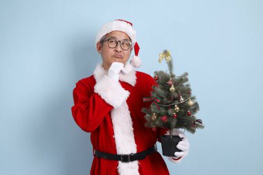 Noel Baba şapkalı Asyalı Noel Baba mavi arka planda izole edilmiş bir Noel ağacı tutuyor..