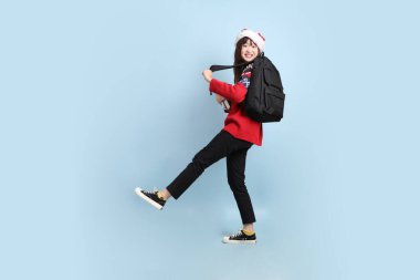 Asyalı bir kız öğrenci, Noel Baba şapkası takmış ve mavi arka planda izole edilmiş bir kitap tutuyor. Noel Baba.