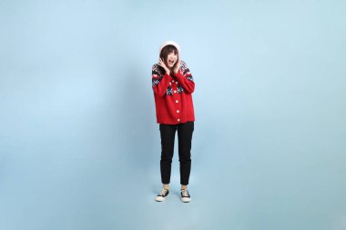 Asyalı bir kadın Noel ya da kışlık kıyafetler giyip Noel Baba şapkası takıyor ve sürpriz bir jest yapıyor mavi arka planda. Noel Baba.