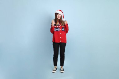 Noel ya da kışlık kıyafetler giyen Asyalı bir kadın Noel Baba şapkası takıyor. Heyecanlı, mavi arka planda izole edilmiş bir şekilde kutluyor. Noel Baba.