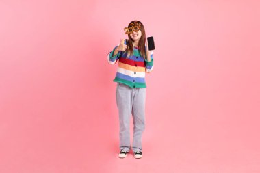 2025 numaralı renkli kış kıyafetleri giyen Asyalı kadın. Yeni yıl gözlüğü. Pembe arka planda izole edilmiş cep telefonu el kol hareketi..