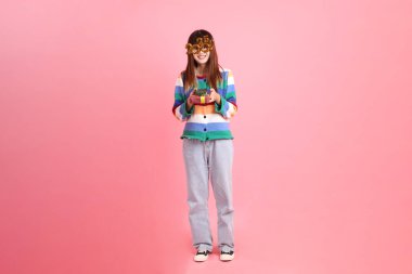 2025 numarayla renkli kışlık kıyafetler giyen Asyalı bir kadın. Yeni yıl gözlüğü. Pembe arka planda saklanmış hediye kutusu jesti..