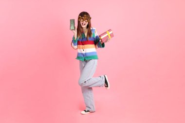 2025 numarayla renkli kışlık kıyafetler giyen Asyalı bir kadın. Yeni yıl gözlüğü. Pembe arka planda saklanmış hediye kutusu jesti..