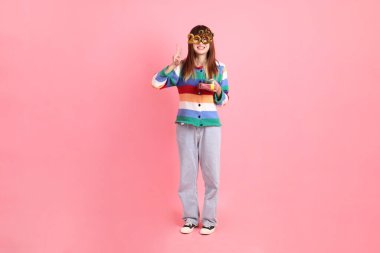 2025 numarayla renkli kışlık kıyafetler giyen Asyalı bir kadın. Yeni yıl gözlüğü. Pembe arka planda saklanmış hediye kutusu jesti..