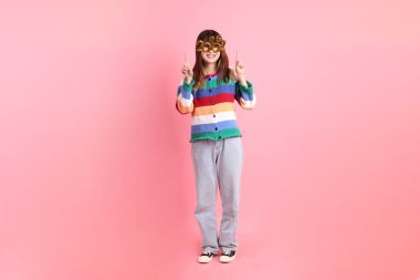 2025 numaralı renkli kış kıyafetleri giyen Asyalı kadın. Yeni yıl gözlüğü. Pembe arka planda izole edilmiş bir işaret..