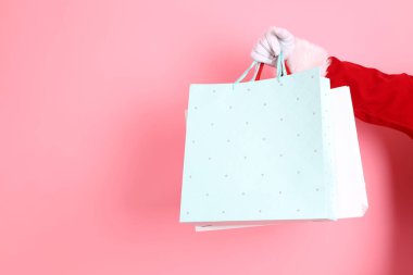 Mutlu Noeller, kışı kutlayan kıdemli adam elinde renkli alışveriş çantaları tutuyor, pembe arka planda Noel Baba kostümü giyiyor. Mutlu Noeller.