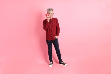Mutlu Noeller, resmi ve sıradan kırmızı tişörtlü Asyalı kıdemli adam. Elinde hediye kutusu olan, pembe arka planda izole edilmiş..
