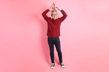 Mutlu Noeller, resmi ve sıradan kırmızı tişörtlü Asyalı kıdemli adam. Elinde hediye kutusu olan, pembe arka planda izole edilmiş..