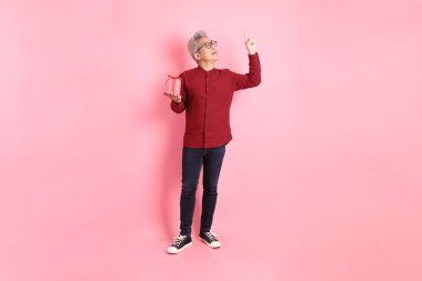 Mutlu Noeller, resmi ve günlük kırmızı gömlekli Asyalı kıdemli adam. Hediye kutusunu tutarken pembe arka planda izole edilmiş bir şekilde işaret ediyor..