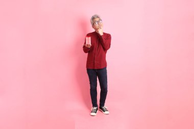 Mutlu Noeller, resmi ve günlük kırmızı tişörtlü Asyalı kıdemli adam. Düşünürken hediye kutusunu tutarken pembe bir arka planda izole edilmiş..