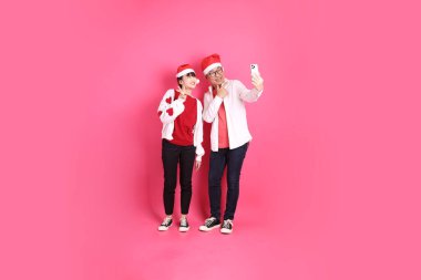 Mutlu Noeller, Asyalı son sınıf öğrencisi ve genç bayan Noel Baba şapkası takın ve pembe arka planda izole edilmiş Holding cep telefonu jesti ile beyaz kalp şeklinde gömlek giyin..