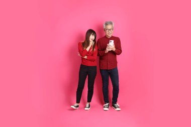 Çin Yeni Yılı, Asyalı son sınıf öğrencisi erkek ve kadın Noel Baba şapkası takıyor, pembe arka planda izole edilmiş cep telefonu jesti ile günlük kırmızı elbiseler giyiyor..