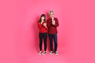 Çin Yeni Yılı, Asyalı son sınıf öğrencisi erkek ve kadın Noel Baba şapkası takıyor, pembe arka planda izole edilmiş cep telefonu jesti ile günlük kırmızı elbiseler giyiyor..