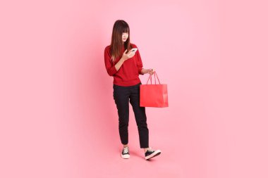 Mutlu Çin Yeni Yılı, pembe arka planda izole edilmiş el çantası ve cep telefonu jesti ile kırmızı günlük Asyalı kadın tişörtü. Gen Z
