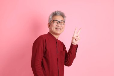 Mutlu Çin Yeni Yılı, resmi ve sıradan kırmızı tişörtlü Asyalı kıdemli barış işareti, V işareti, pembe arka planda iki parmak izole edilmiş..