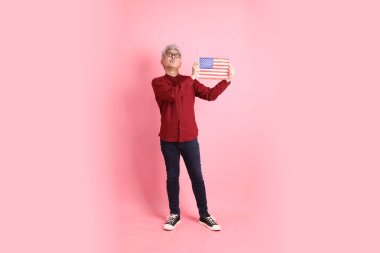 ABD, Amerika ya da Amerika Birleşik Devletleri ya da Amerika bayrağını pembe arka planda tutan resmi ve resmi kırmızı tişörtlü Asyalı kıdemli adam..