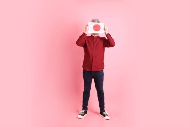 Pembe arka planda Japon ya da Japon bayrağı taşıyan resmi ve sıradan kırmızı tişörtlü Asyalı kıdemli bir adam..