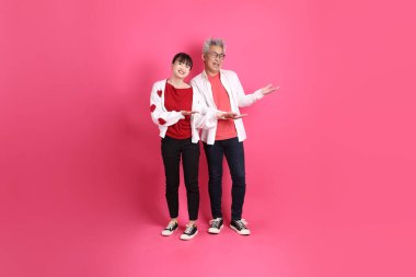 Sevgililer Günü 'nde, Asyalı son sınıf öğrencisi ve genç kadın pembe arka planda izole edilmiş işaret eden beyaz kalp şeklinde gömlek ve günlük kırmızı kıyafetler giyerler..