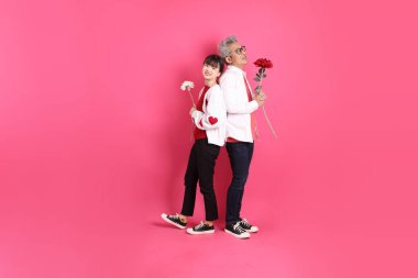 Sevgililer Günü 'nde, Asyalı son sınıf öğrencisi ve genç kadın pembe arka planda saklanmış çiçekleri tutan beyaz kalp şeklinde gömlek ve günlük kırmızı kıyafetler giyerler..