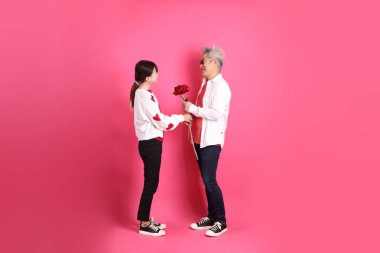 Sevgililer Günü 'nde, Asyalı son sınıf öğrencisi ve genç kadın pembe arka planda saklanmış çiçekleri tutan beyaz kalp şeklinde gömlek ve günlük kırmızı kıyafetler giyerler..