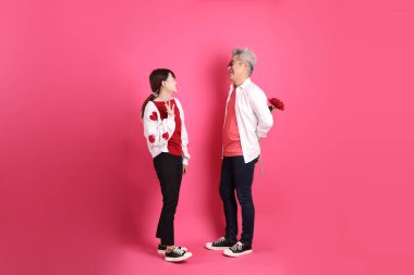 Sevgililer Günü 'nde, Asyalı son sınıf öğrencisi ve genç kadın pembe arka planda saklanmış çiçekleri tutan beyaz kalp şeklinde gömlek ve günlük kırmızı kıyafetler giyerler..