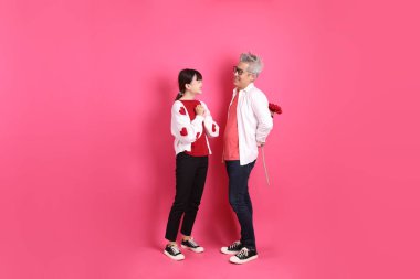 Sevgililer Günü 'nde, Asyalı son sınıf öğrencisi ve genç kadın pembe arka planda saklanmış çiçekleri tutan beyaz kalp şeklinde gömlek ve günlük kırmızı kıyafetler giyerler..