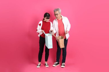 Sevgililer Günü, Asyalı son sınıf öğrencisi ve genç kadın pembe arka planda izole edilmiş alışveriş çantası jesti içeren beyaz kalp şeklinde gömlek ve günlük kırmızı kıyafetler giyerler..