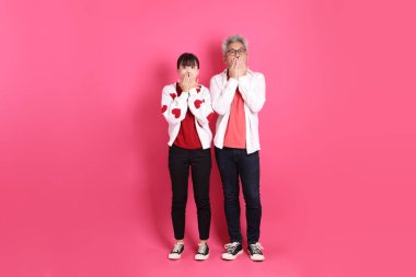 Sevgililer Günü 'nde, Asyalı son sınıf öğrencisi ve genç bir kadın pembe arka planda izole edilmiş, ağzı kapalı, sessiz bir jest içeren, beyaz kalp şeklinde gömlek ve günlük kırmızı kıyafetler giyerler..