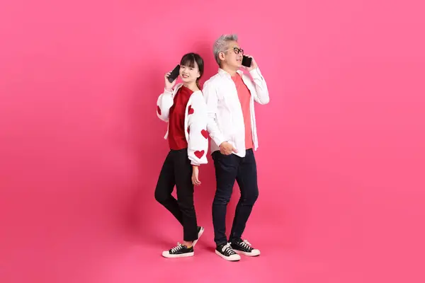 Sevgililer Günü 'nde, Asyalı son sınıf öğrencisi ve genç kadın pembe arka planda izole edilmiş Holding cep telefonu jesti içeren beyaz kalp şeklinde gömlekli sıradan kırmızı kıyafetler giyerler..