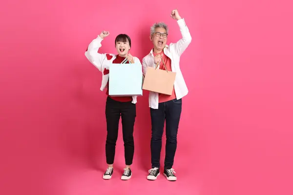 Sevgililer Günü, Asyalı son sınıf öğrencisi ve genç kadın pembe arka planda izole edilmiş alışveriş çantası jesti içeren beyaz kalp şeklinde gömlek ve günlük kırmızı kıyafetler giyerler..