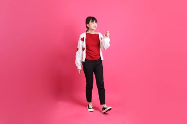 Sevgililer Günü, kırmızı tişörtlü Asyalı bir kadın ya da günlük kıyafetler, pembe arka planda izole edilmiş yürüyen bir jestle kırmızı kalp şeklinde gömlek. Gen Z