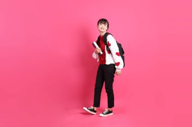 Kırmızı tişörtlü Asyalı kız öğrenci ya da günlük kıyafetler, kırmızı kalp şeklinde gömlek okul çantası ve kitap taşıyor ve pembe arka planda tek başına ayakta durup gülümsüyor. Gen Z