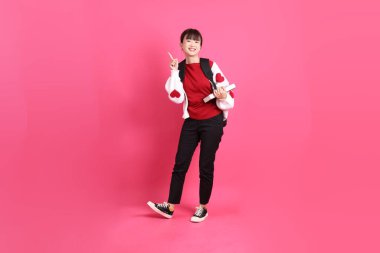 Kırmızı tişörtlü Asyalı kız öğrenci ya da günlük kıyafetler, kırmızı kalp şeklinde gömlek okul çantası ve kitap taşıyor pembe arka planda izole edilmiş bir işaret işareti var. Gen Z