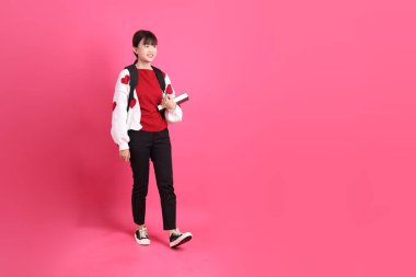 Kırmızı tişörtlü Asyalı kız öğrenci ya da günlük kıyafetler, kırmızı kalp şeklinde gömlek okul çantası ve kitabı pembe arka planda izole edilmiş yürüyüş jestleriyle taşıyor. Gen Z
